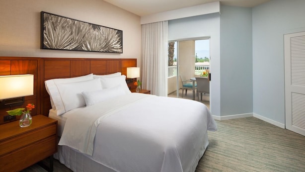 A hotel room with a large bed, a nightstand with a lamp, a vase with flowers, and a view of the outdoors through a sliding door. - The Westin Mission Hills Resort Villas, Palm Springs (Rancho Mirage)