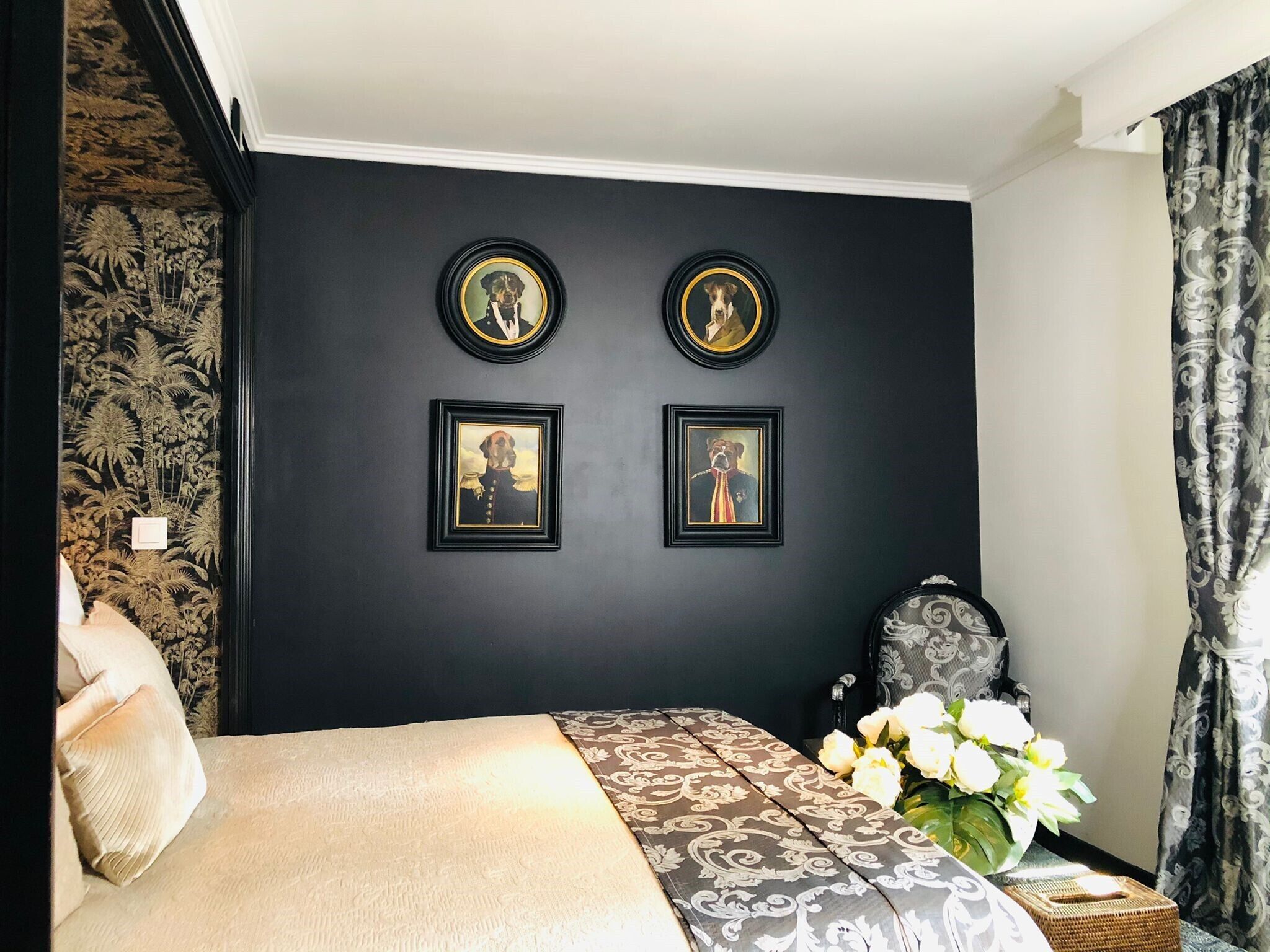 A bedroom with a bed, a wall with framed pictures, a patterned table, and a vase of flowers.