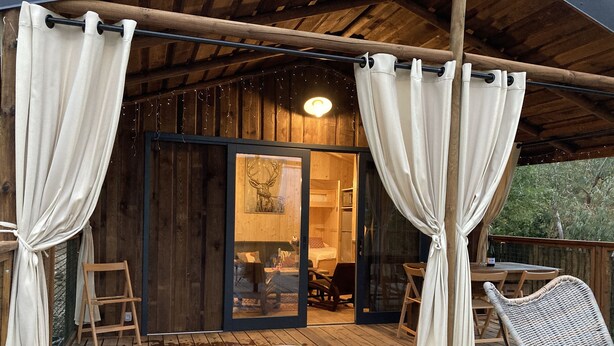 A wooden deck with a hot tub, white curtains, and a glass door leading to a room with a bed and a table. - Cabane Insolite et Nature Avec Jacuzzi et Piscine Prés Carcassonne Occitanie (VILLARZEL DU RAZES)