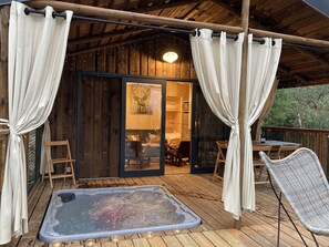 A wooden deck with a hot tub, white curtains, and a glass door leading to a room with a bed and a table. - Cabane Insolite et Nature Avec Jacuzzi et Piscine Prés Carcassonne Occitanie (VILLARZEL DU RAZES)