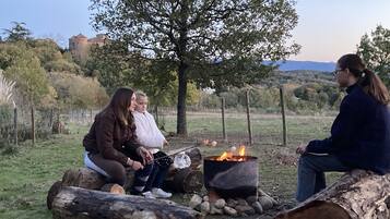 A group of people sitting around a campfire in a grassy outdoor area with trees and a castle in the background.