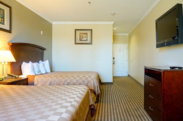 A hotel room with two beds, a desk with a lamp, a dresser, a television, and a framed picture on the wall. - Harbor Inn And Suites (Garden Grove)