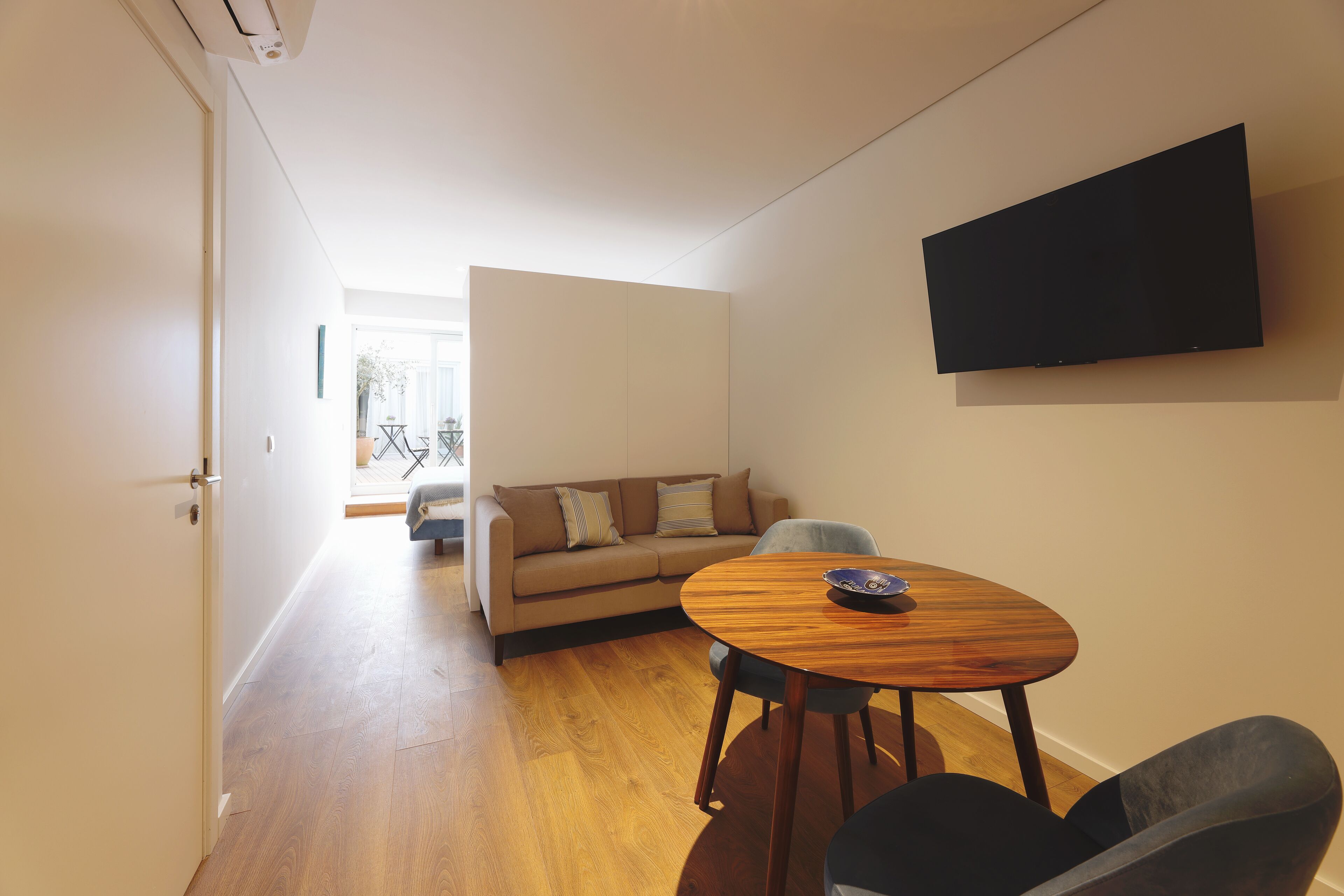 Een moderne woonkamer met houten vloer, een beige bank, een ronde houten tafel en een flatscreen televisie aan de muur.