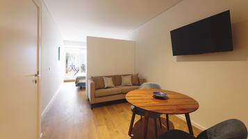 Een moderne woonkamer met houten vloer, een beige bank, een ronde houten tafel en een flatscreen televisie aan de muur.