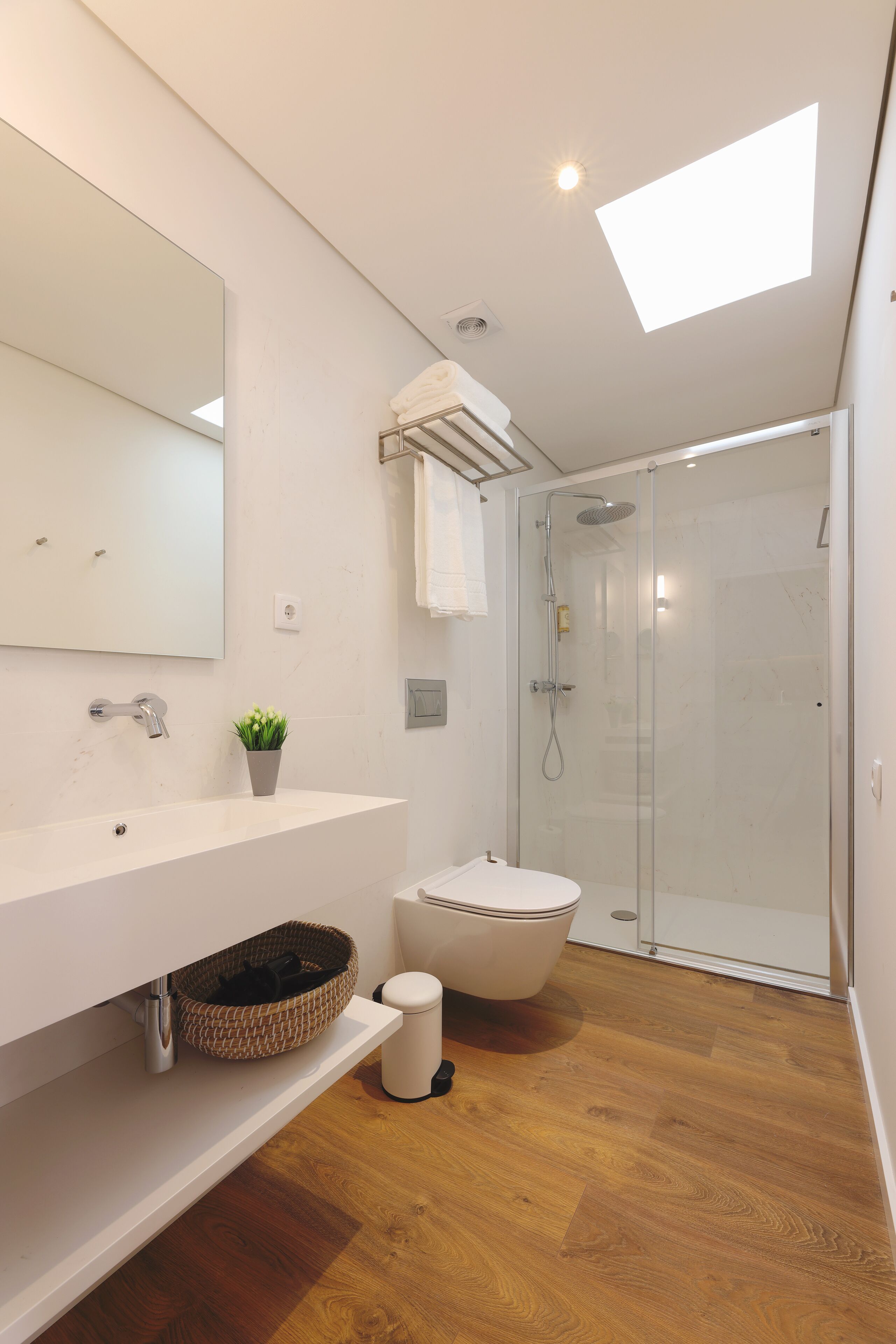 Une salle de bain moderne avec un puits de lumière, un grand miroir, un lavabo blanc, un espace douche et un plancher en bois.