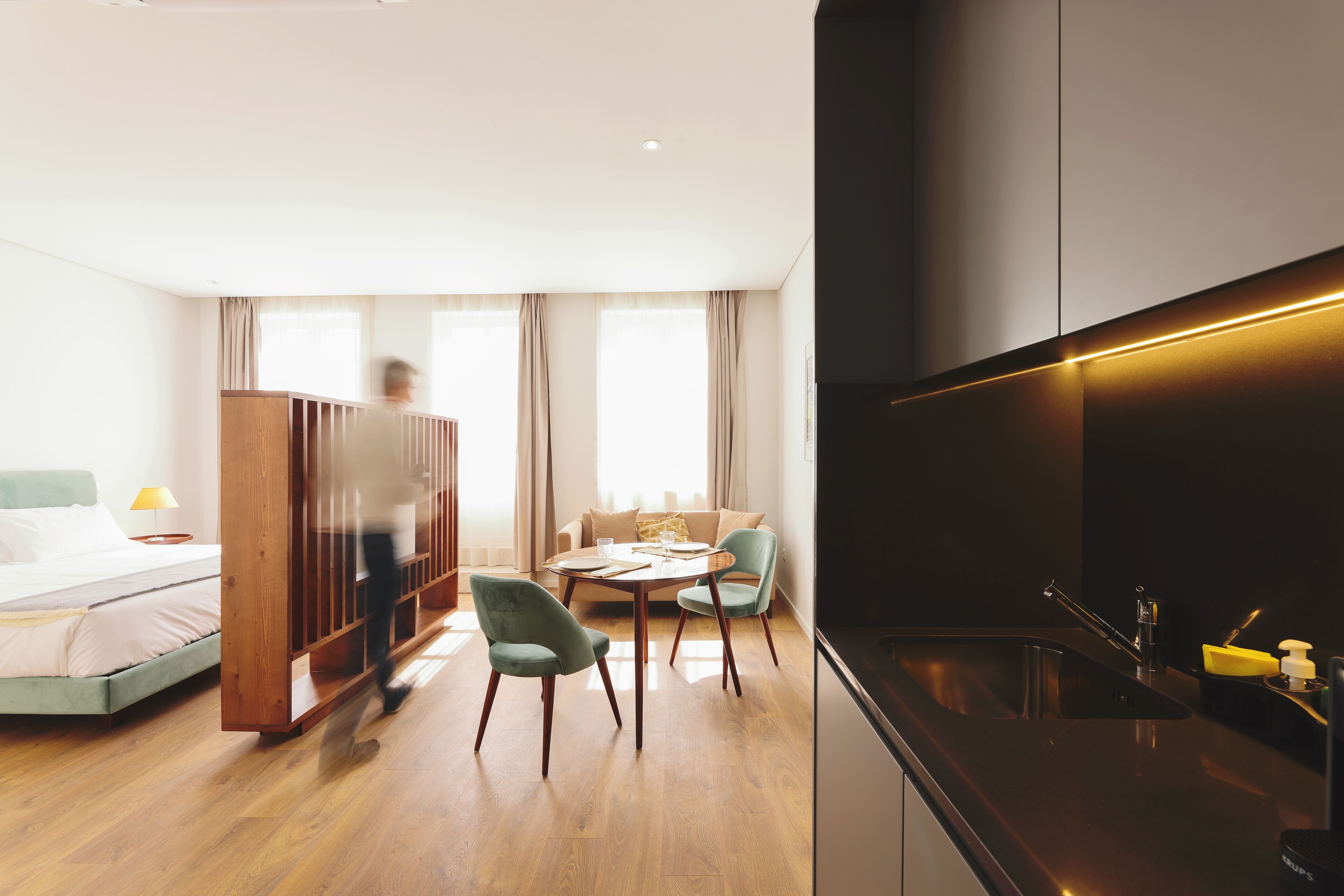 A modern hotel room with a bed, a wooden wardrobe, a dining table with chairs, a sofa, and a kitchen area with a sink and countertop.