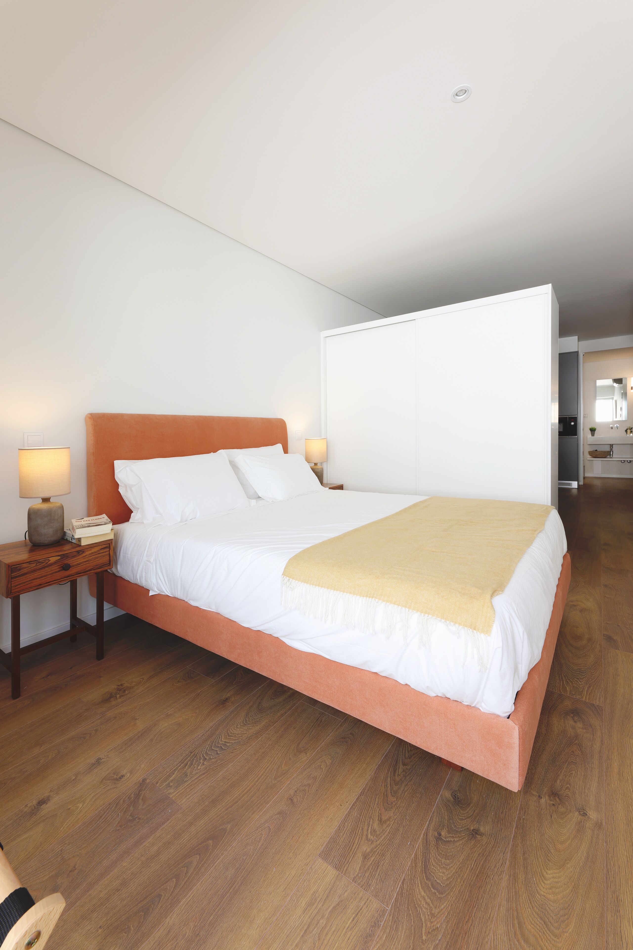 Een slaapkamer met een houten vloer, een bed met wit beddengoed, een oranje hoofdeinde en bedlampjes.