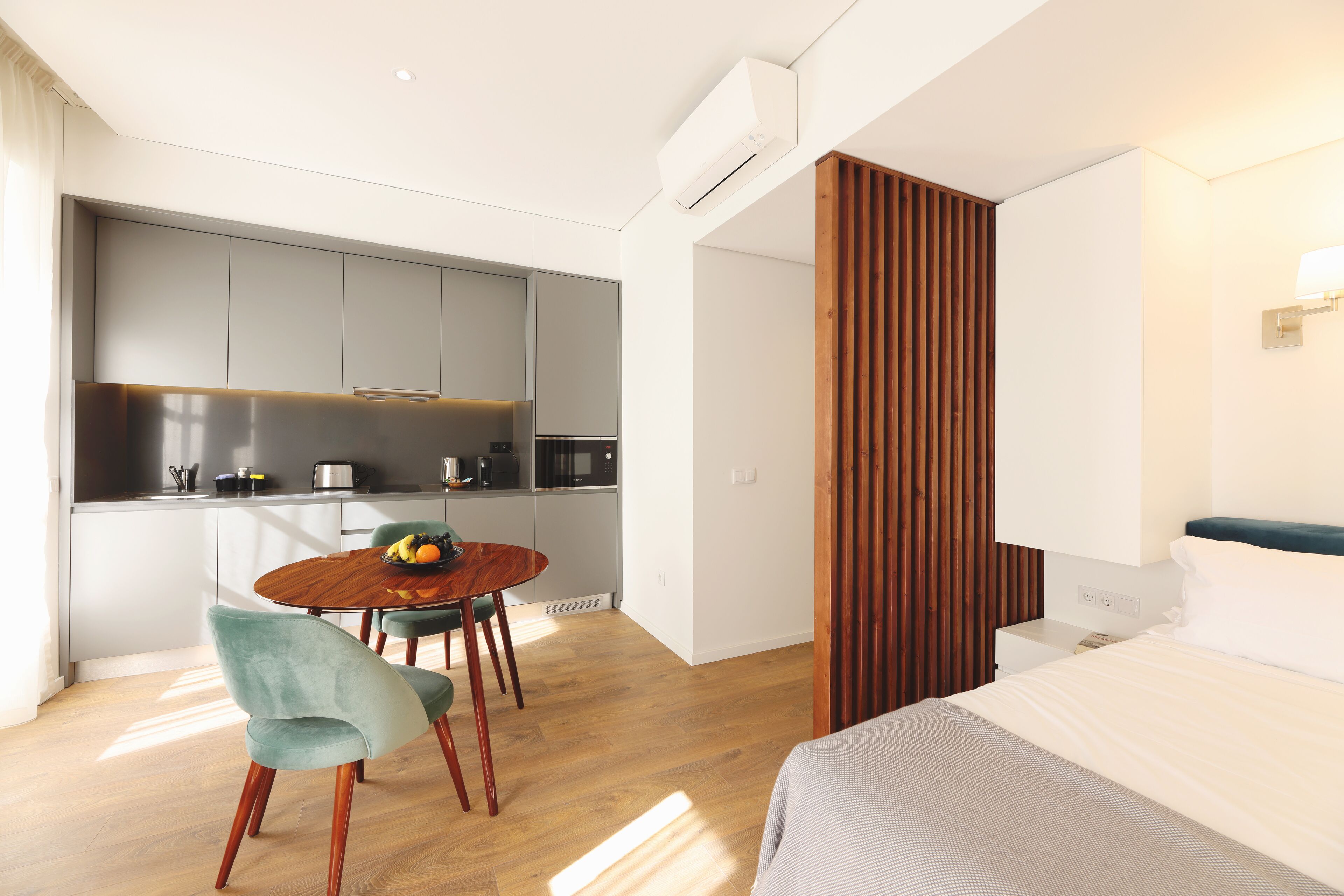 Une chambre d’hôtel moderne avec une table à manger en bois, un lit doté d’une tête de lit blanche et un coin cuisine avec des meubles gris et des appareils électroménagers en acier inoxydable.