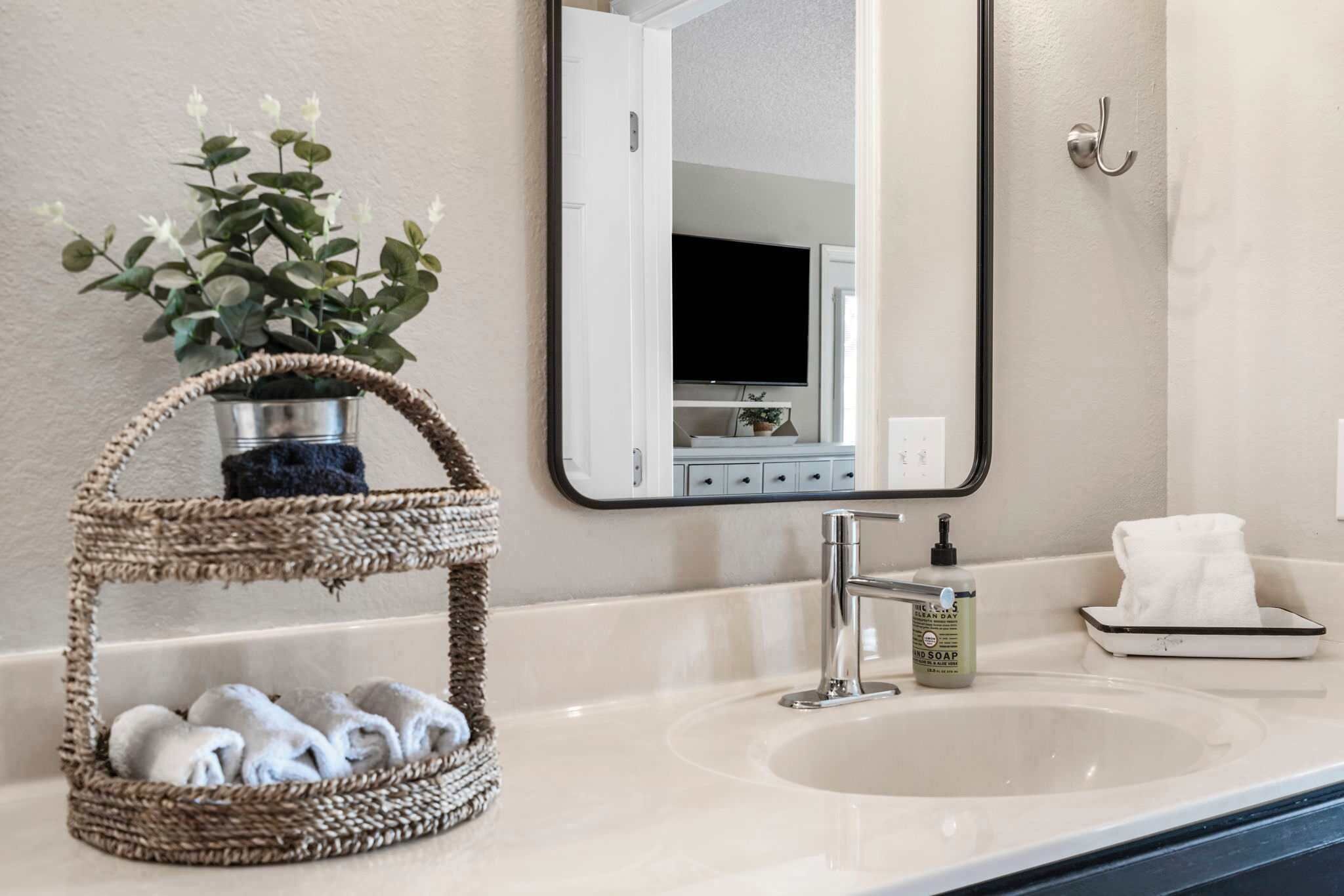 Une salle de bain avec un panier en osier contenant des serviettes, un lavabo, un distributeur de savon et un miroir qui reflète un téléviseur et une porte.