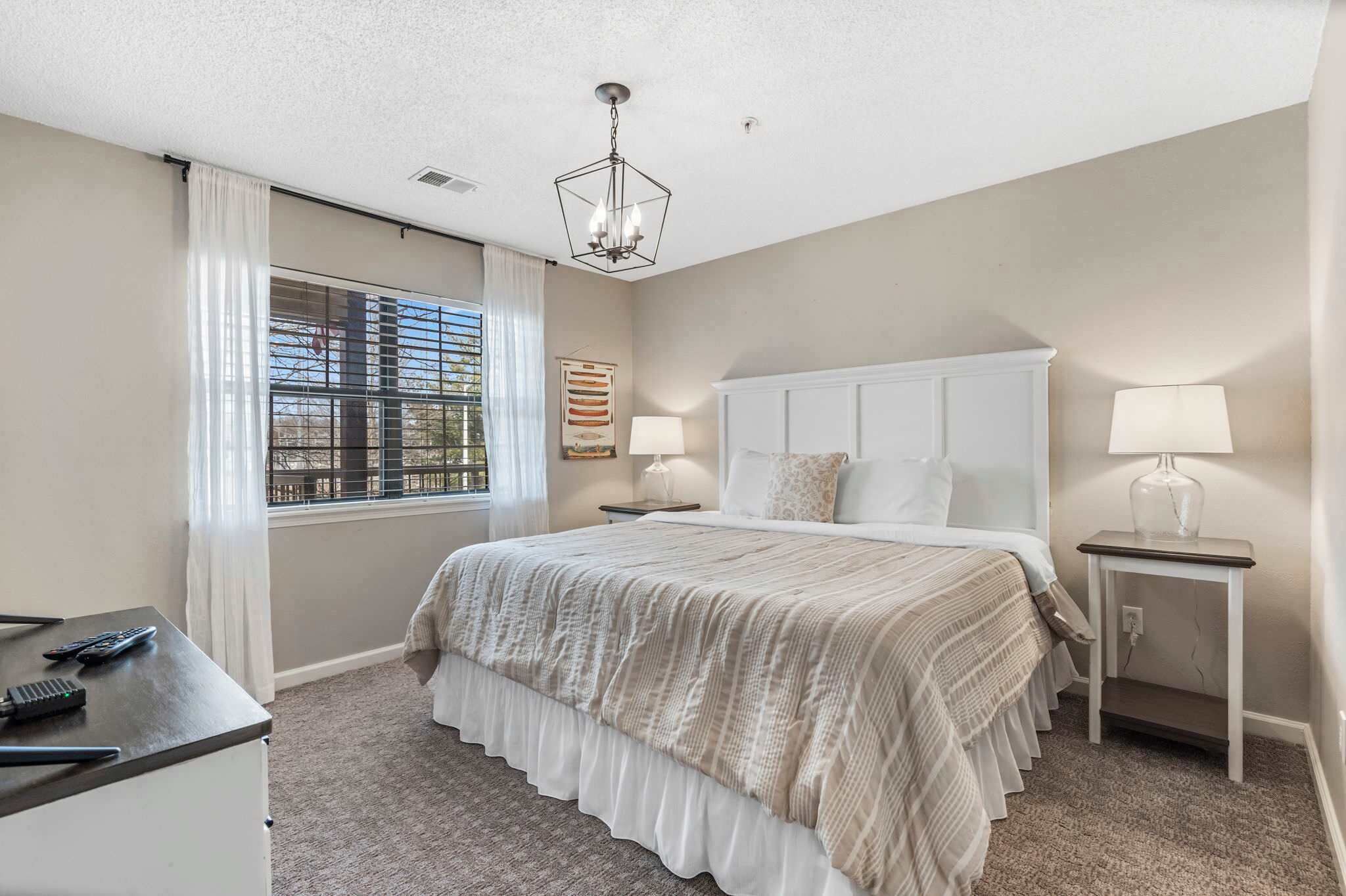 A bedroom with a large bed, a desk, a chair, a window with blinds, a chandelier, and a nightstand with a lamp.
