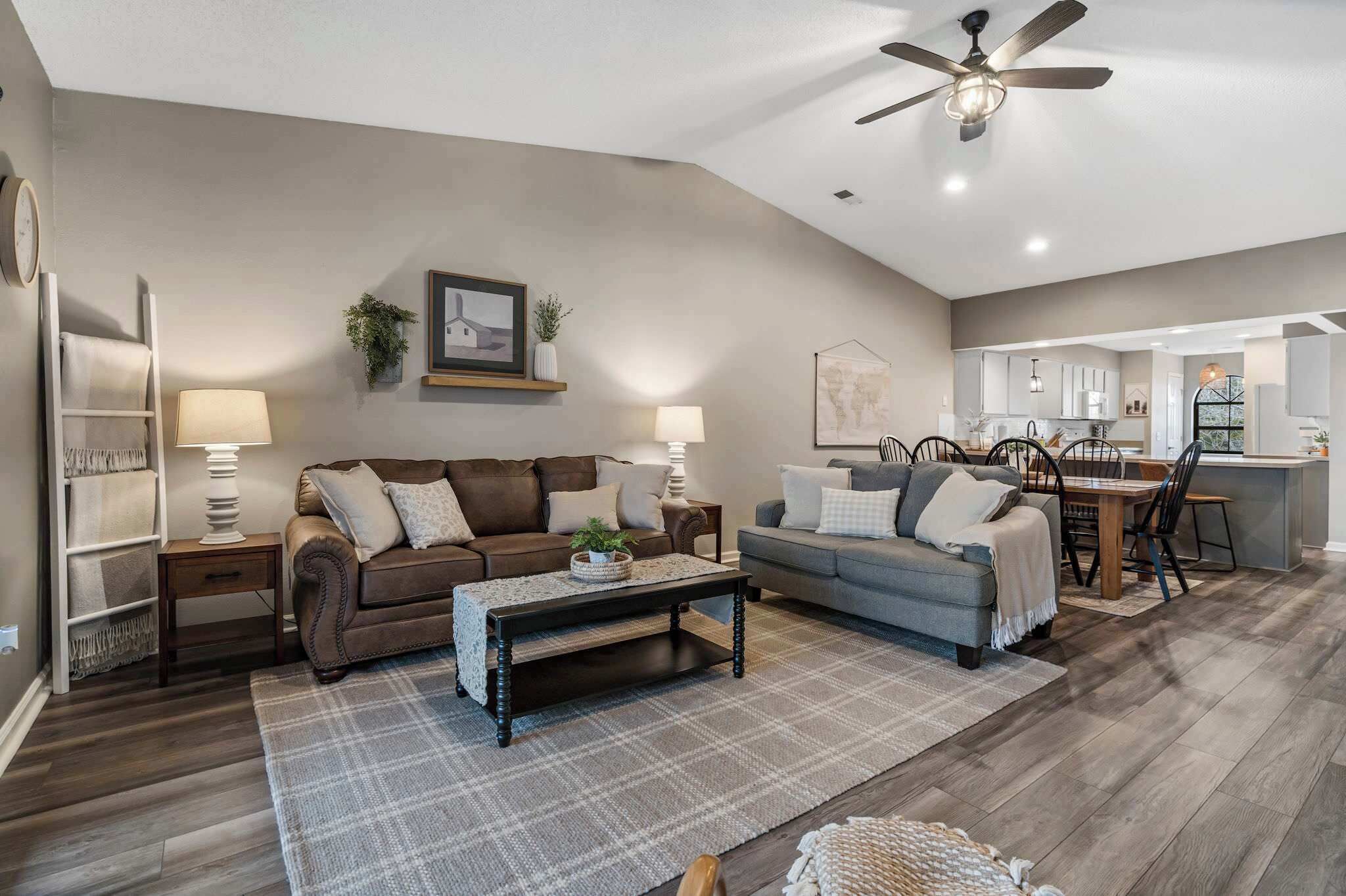 A spacious living area with a brown sofa, a grey sofa, and a wooden coffee table. There is a dining table with chairs, a ceiling fan, and a kitchen area in the background.