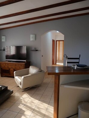 A living room with a TV, sofa, dining table, and a kitchen area in the background. - Appartement cozy en résidence (Libreville)