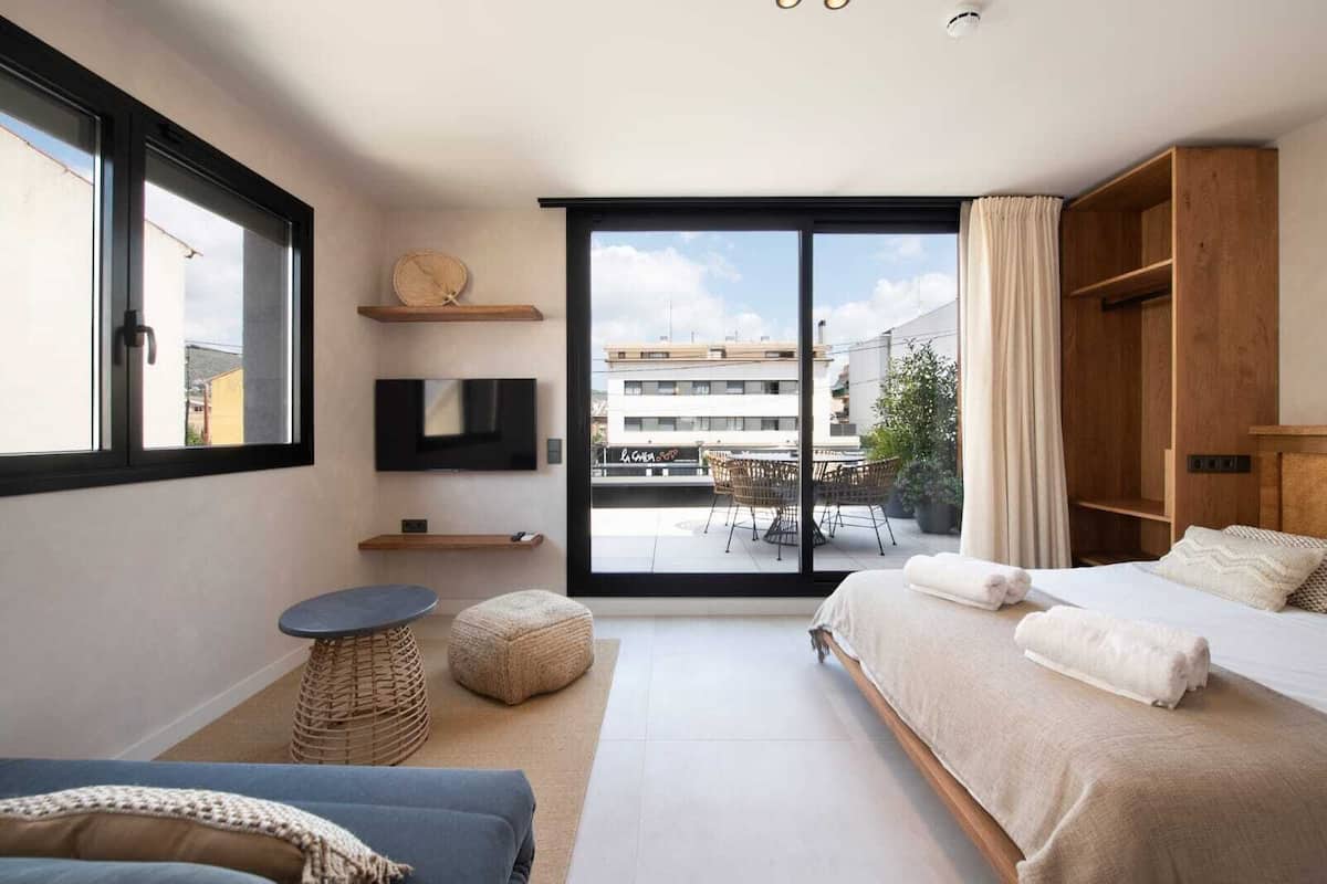 A modern bedroom with a large bed, a TV, a small table, and a view of the city through a sliding door.