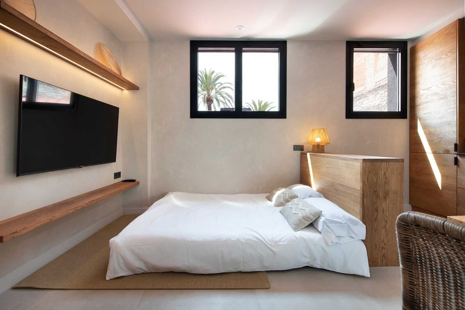 A modern bedroom with a bed, a TV mounted on the wall, and two windows providing natural light.