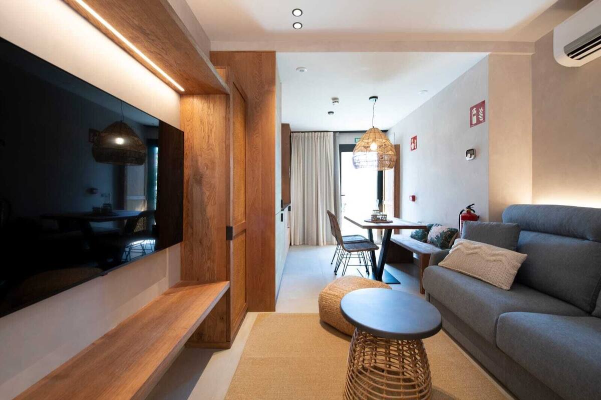 A modern living room with a flat-screen TV, a grey sofa, a wooden bench, and a dining area with a table and chairs.