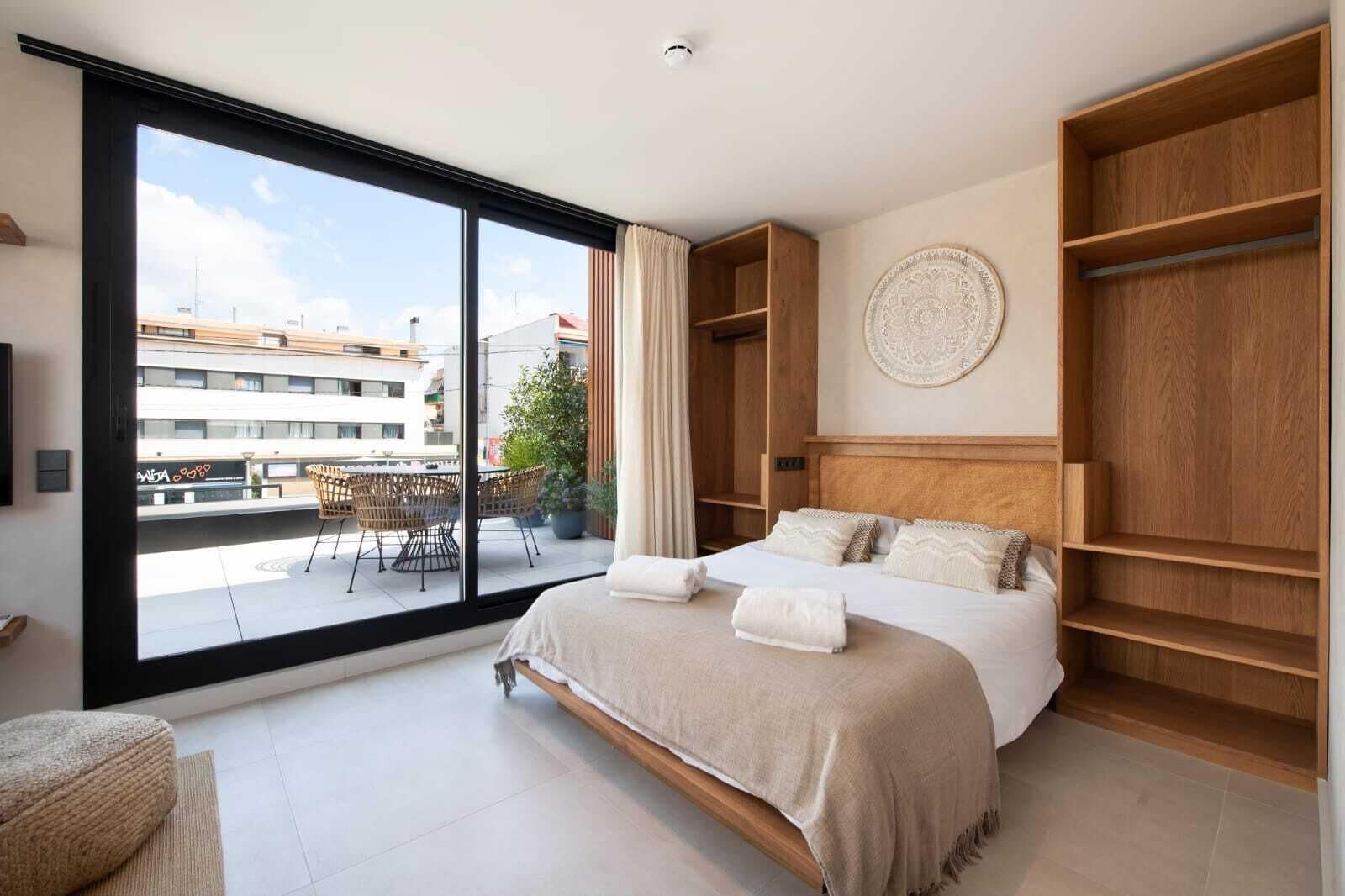 Une chambre moderne dotée d’une grande porte-fenêtre coulissante donnant sur un balcon, d’un lit avec une tête de lit et d’étagères en bois intégrées.