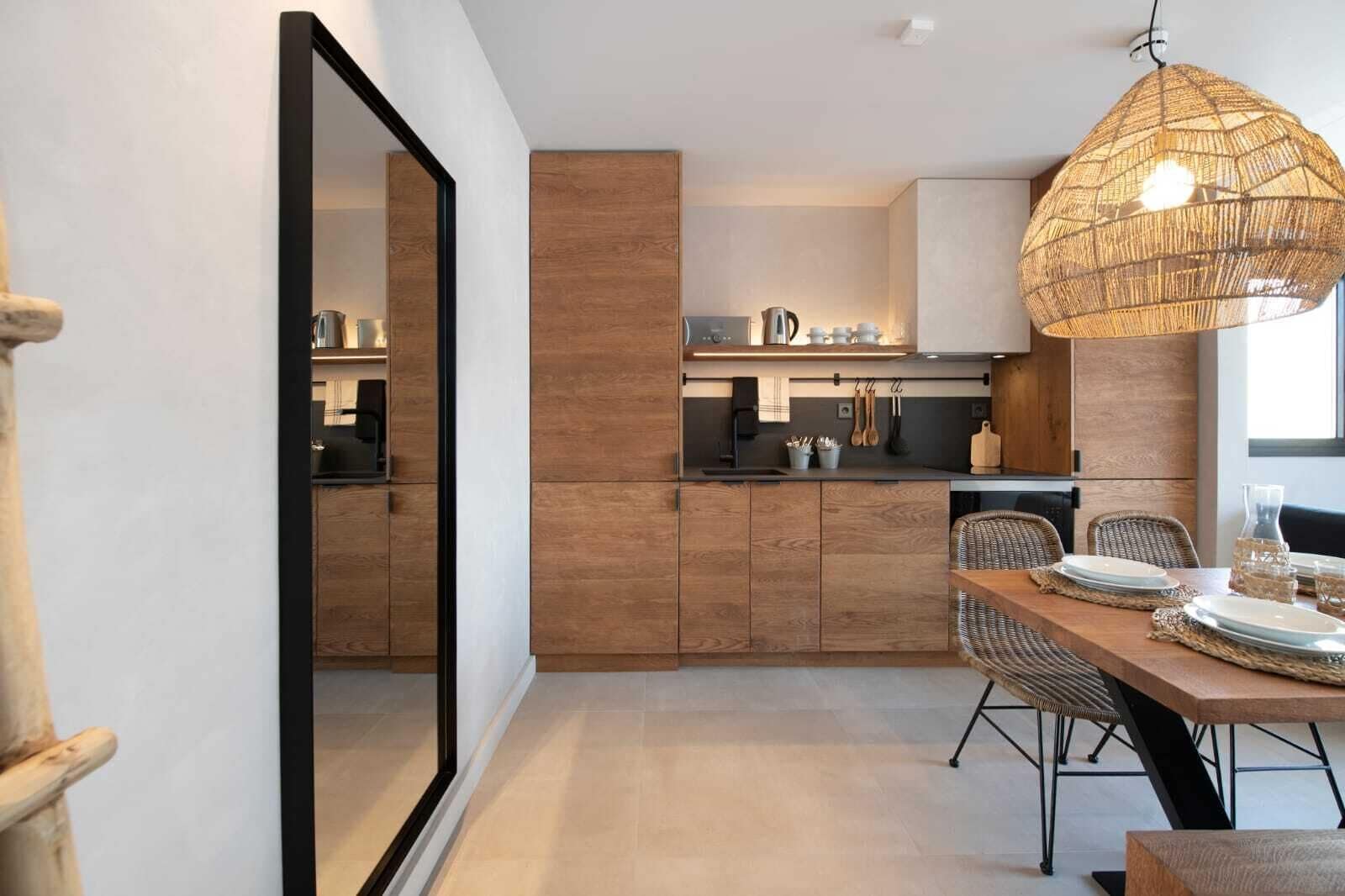 A modern kitchen with wooden cabinets, a dining area with wicker chairs, and a large mirror.