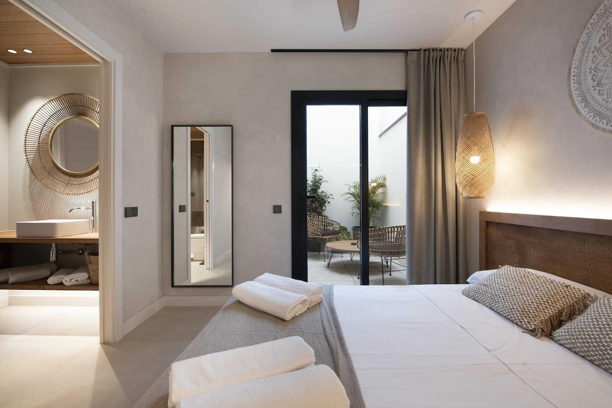 A modern hotel room with a large bed, a bathroom with a round mirror, a balcony with seating, and a view of the outdoors.