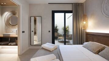 A modern hotel room with a large bed, a bathroom with a round mirror, a balcony with seating, and a view of the outdoors.