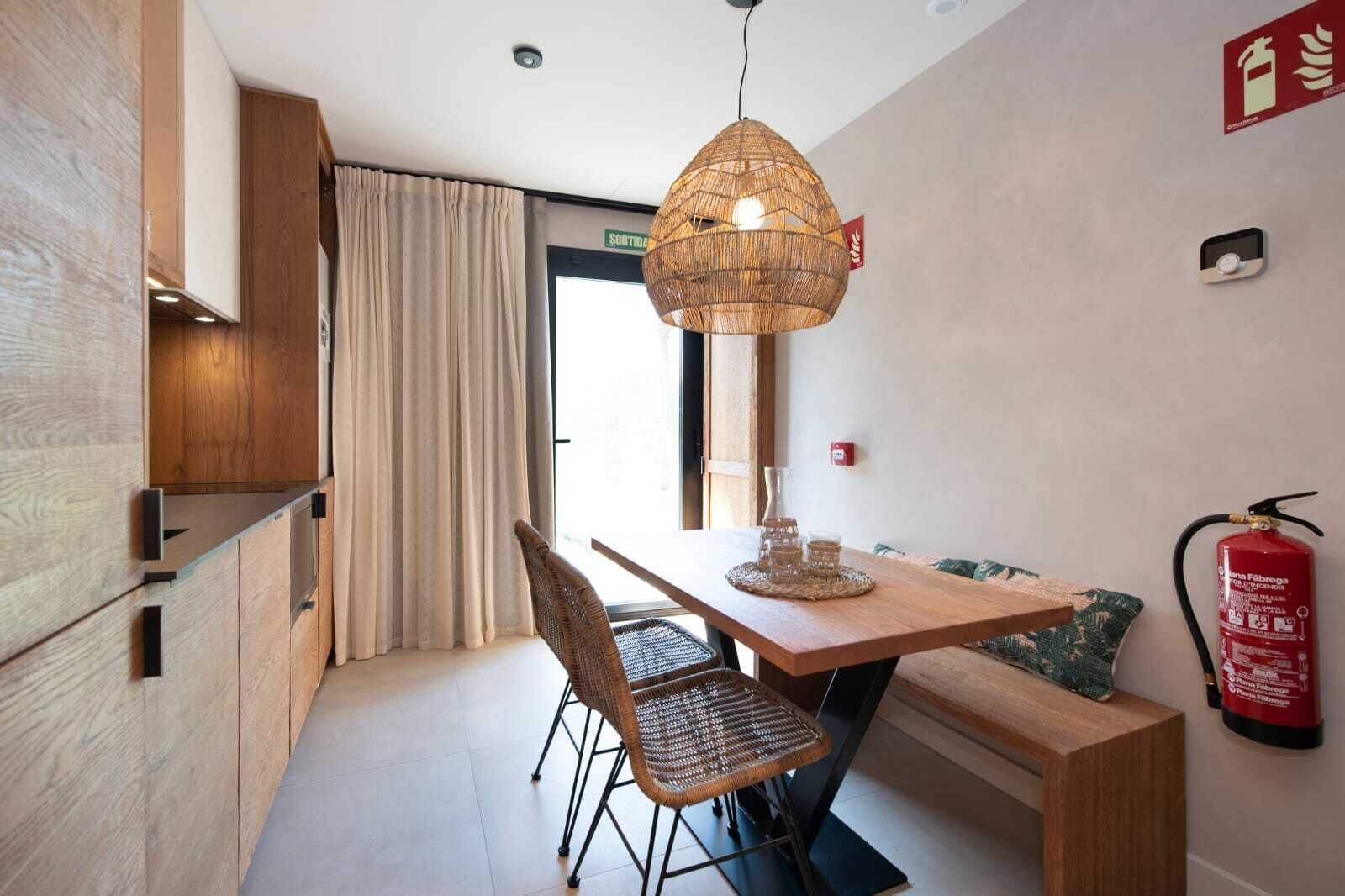 A modern hotel room with a wooden dining table, wicker chairs, a hanging light fixture, and a fire extinguisher on the wall.