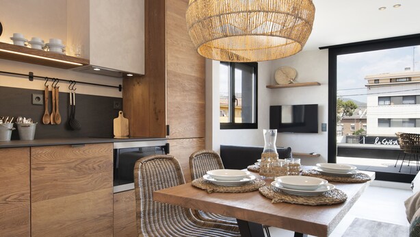 A modern kitchen with wooden cabinets, a dining area with wicker chairs, and a large woven pendant light. - Apartamentos Balisse (Castelldefels)