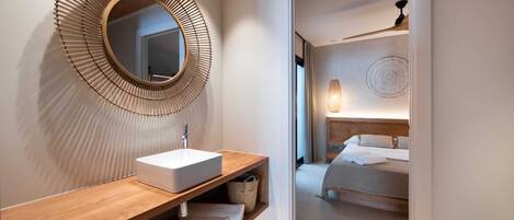 A modern bathroom with a wooden vanity, a large mirror, and a bed visible through an open door.