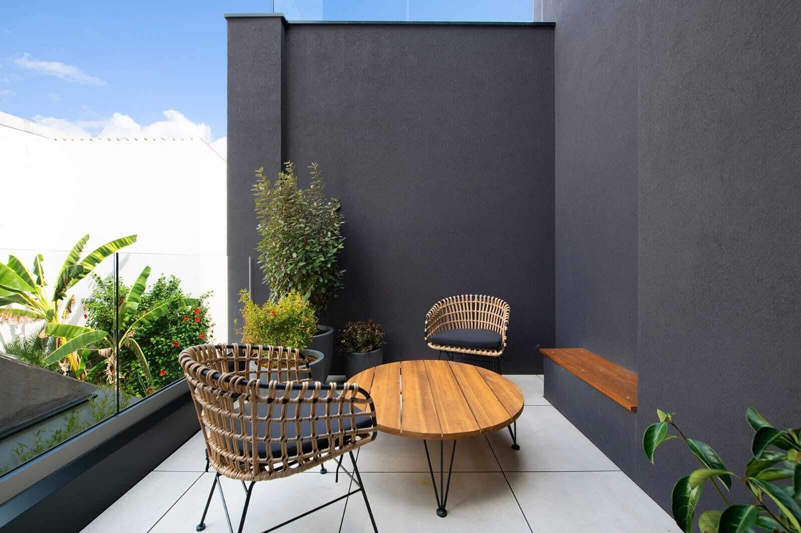 Een moderne zithoek buiten, met rieten stoelen en een houten tafel, omringd door potplanten en een donkere muur.