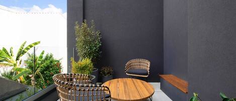 Een moderne zithoek buiten, met rieten stoelen en een houten tafel, omringd door potplanten en een donkere muur.
