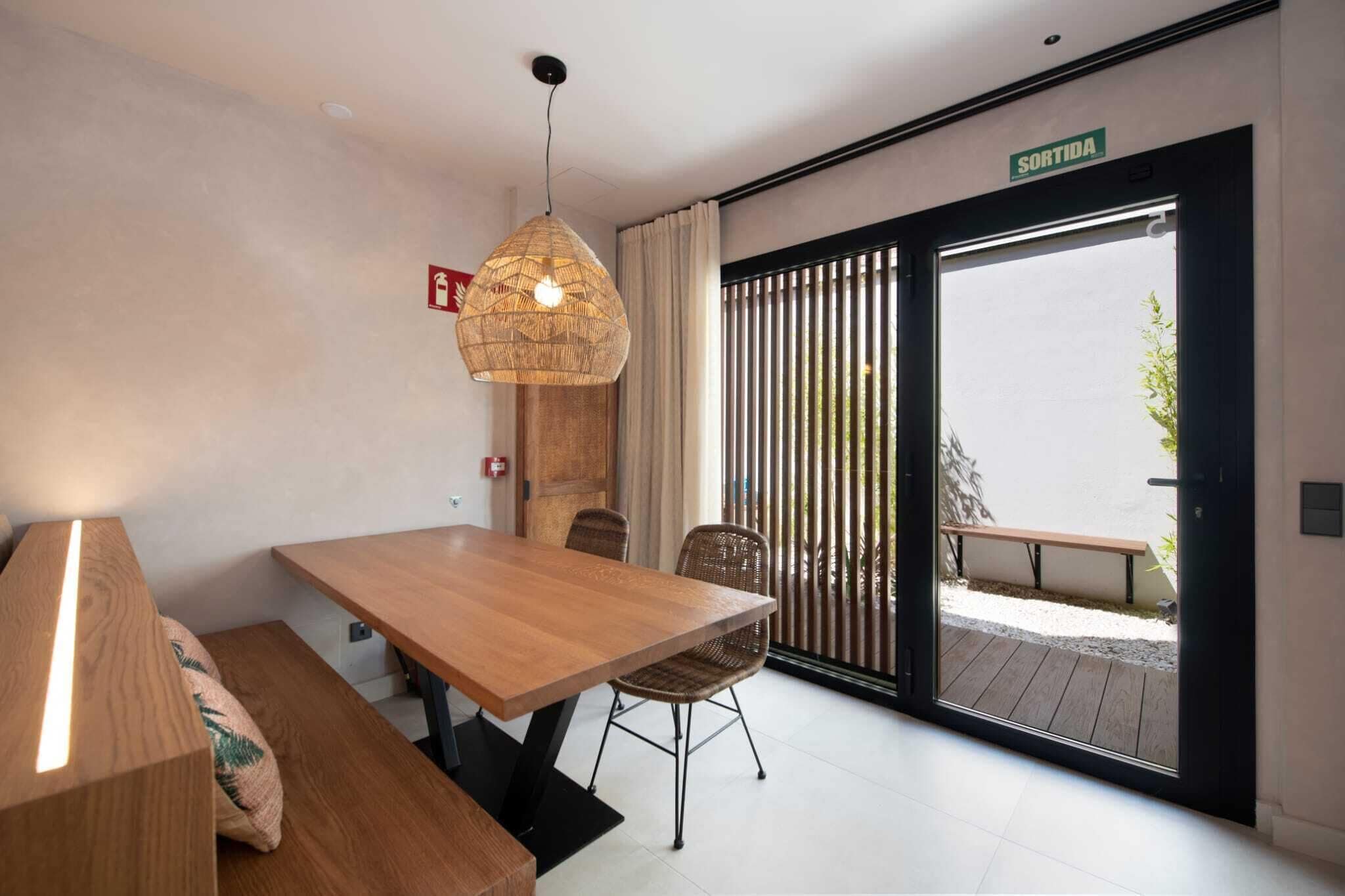 Un comedor moderno con una mesa y sillas de madera, una lámpara colgante y un ventanal corredero que da a un patio exterior.