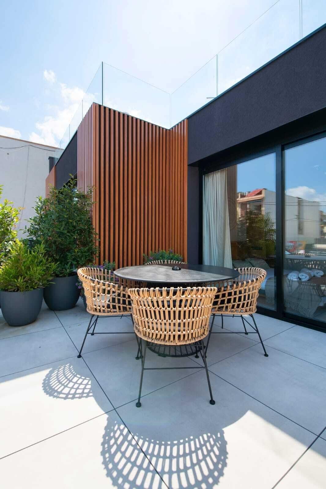 Un patio moderno al aire libre con una mesa redonda y sillas de mimbre, rodeado de plantas en macetas.