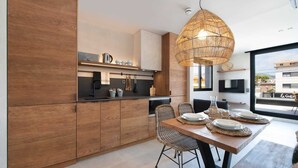 A modern kitchen with wooden cabinets, a dining area with wicker chairs, and a large window offering a view of the city. - Apartamentos Balisse (Castelldefels)