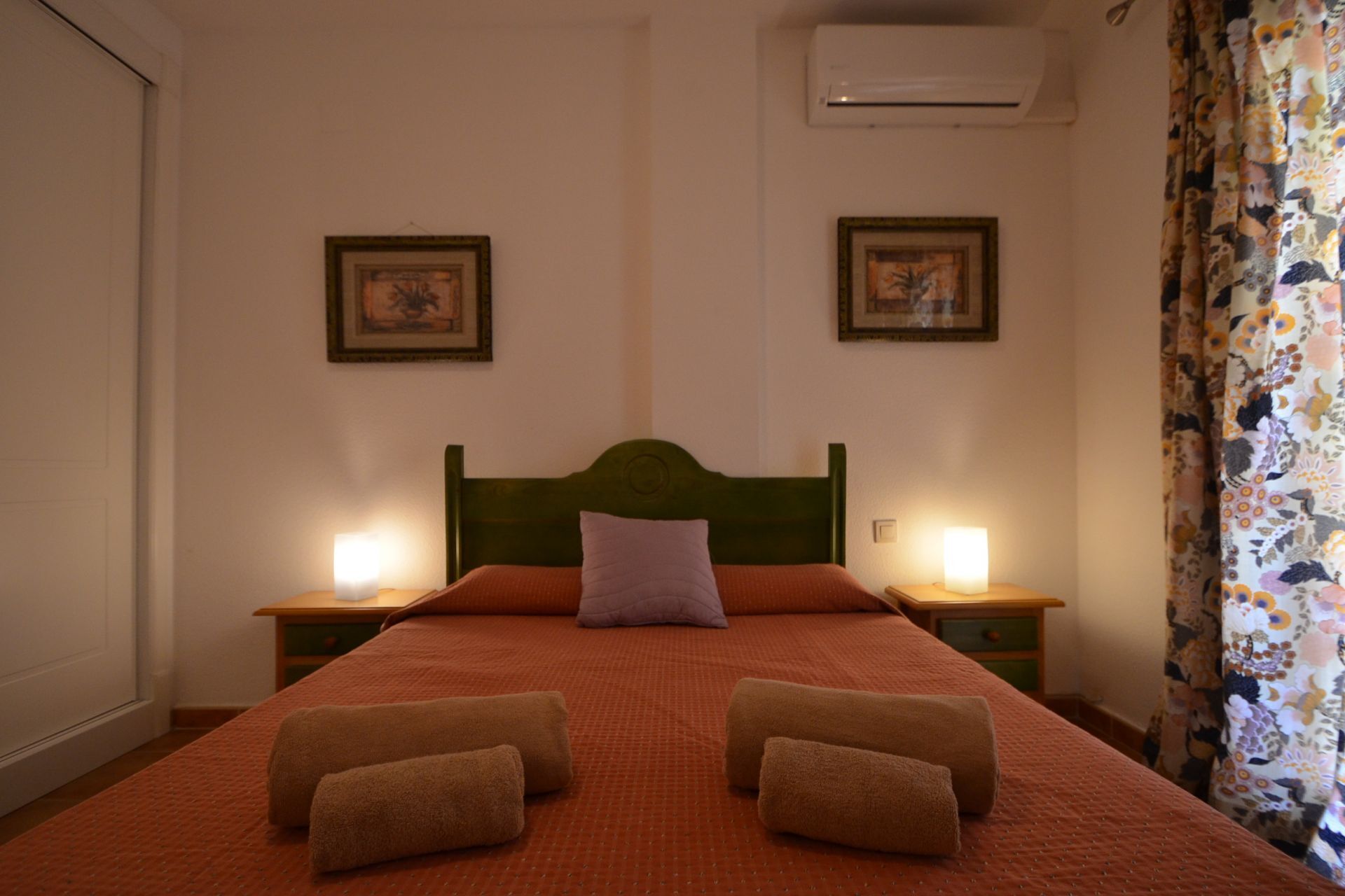 A neatly made bed with two pillows and two rolled towels, two bedside lamps, and two framed pictures on the wall.