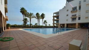 A swimming pool with a tiled deck, surrounded by palm trees and a multi-story building with balconies. - Espigón Poniente 7 - Two Bedroom Apartment, Sleeps 5 (Punta del moral)