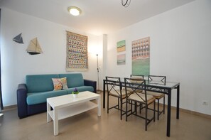 Un espacio de estar con un sofá color turquesa, una mesa de centro blanca, una mesa de comedor con cubierta de vidrio y cuatro sillas, y decoración de pared que incluye un velero y un tapiz colorido.