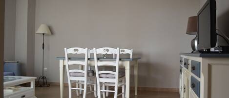 Un coin repas comprenant une table et des chaises, une lampe, un téléviseur posé sur un meuble et une bibliothèque.