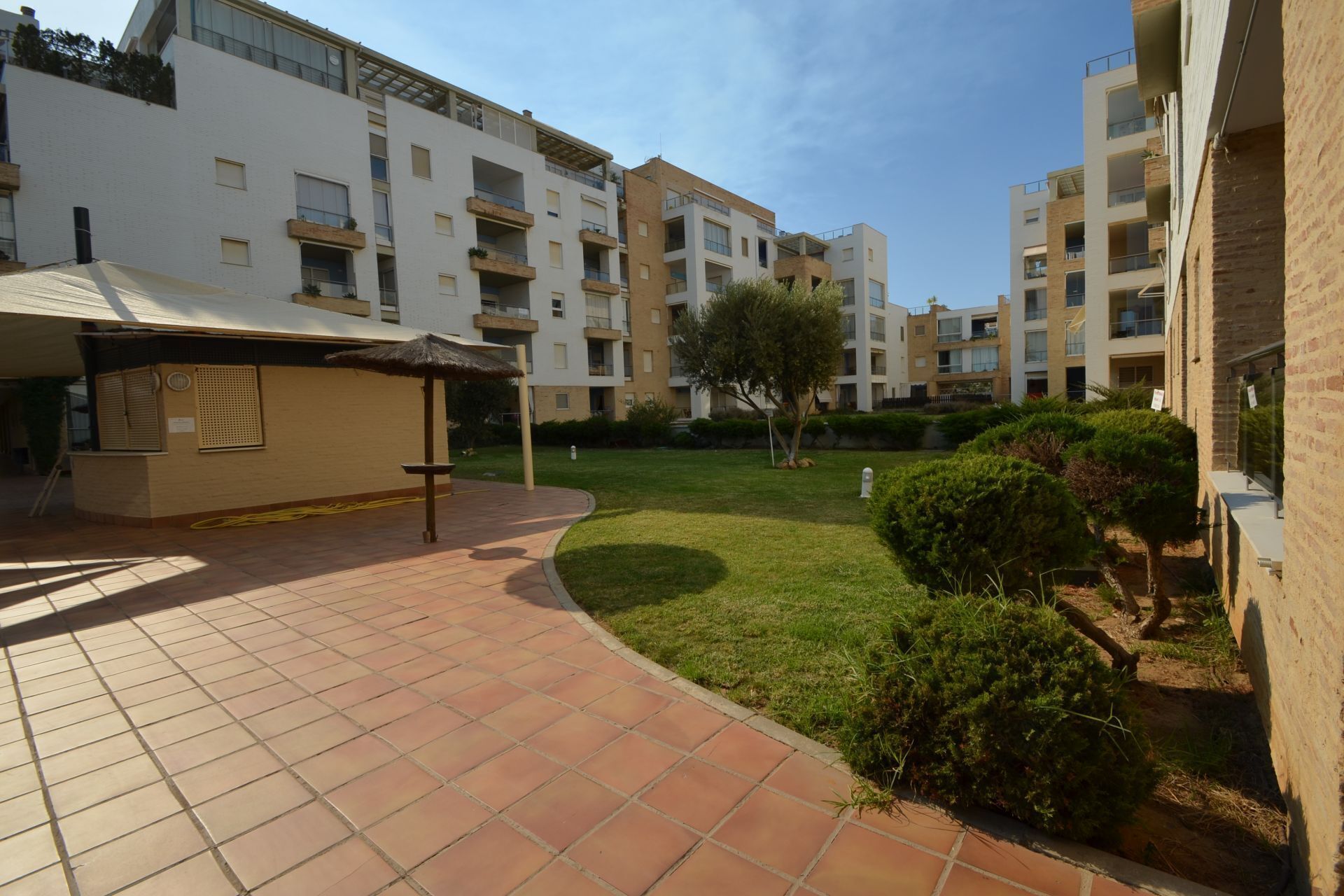 A courtyard with a tiled walkway, a covered seating area, and a grassy lawn with bushes.