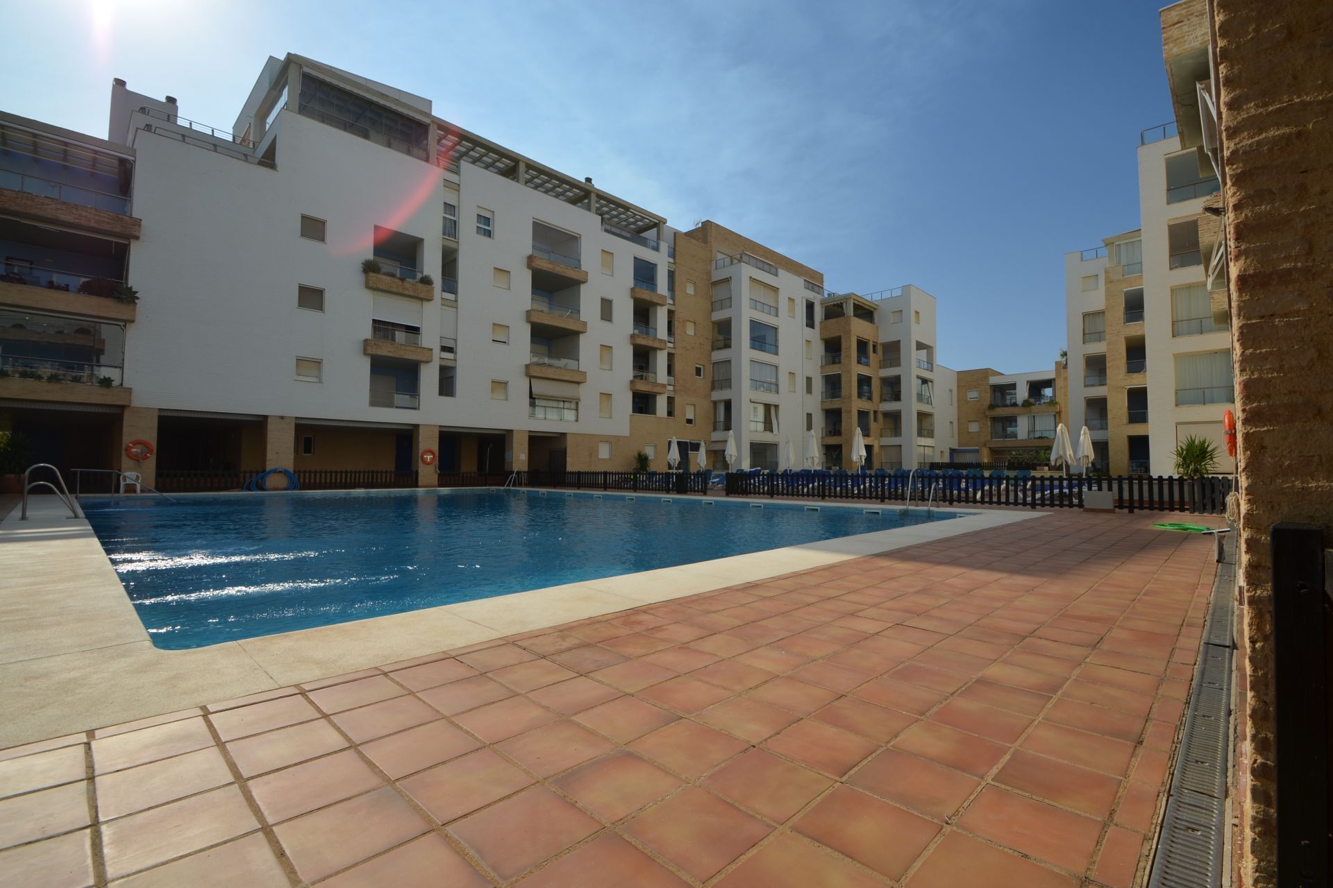 A swimming pool area with a tiled deck, surrounded by multi-story buildings with balconies and windows.
