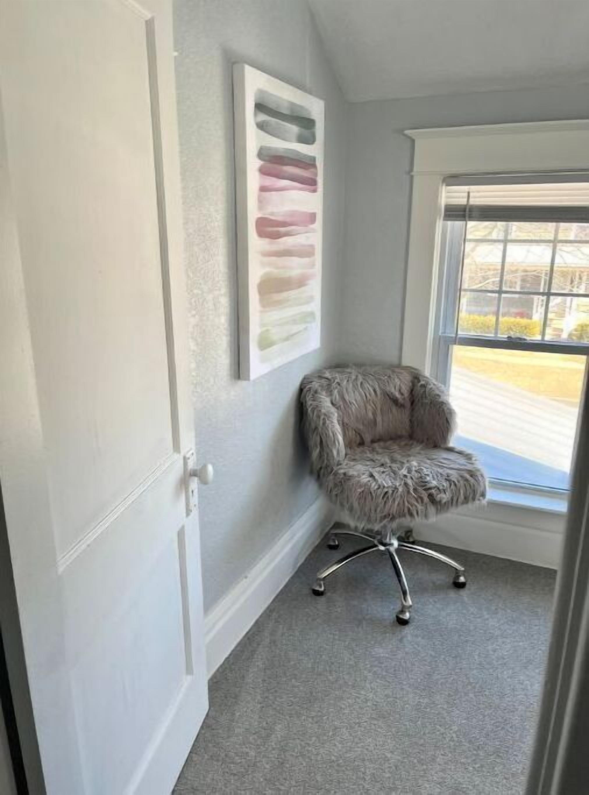 Una habitación con alfombra gris, una butaca gris y mullida, una puerta blanca y un ventanal con vistas a una carretera y árboles.