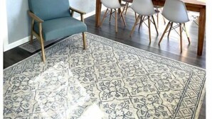 Un coin repas avec une table et des chaises en bois, un fauteuil bleu, un tapis à motifs et une œuvre d’art accrochée au mur.