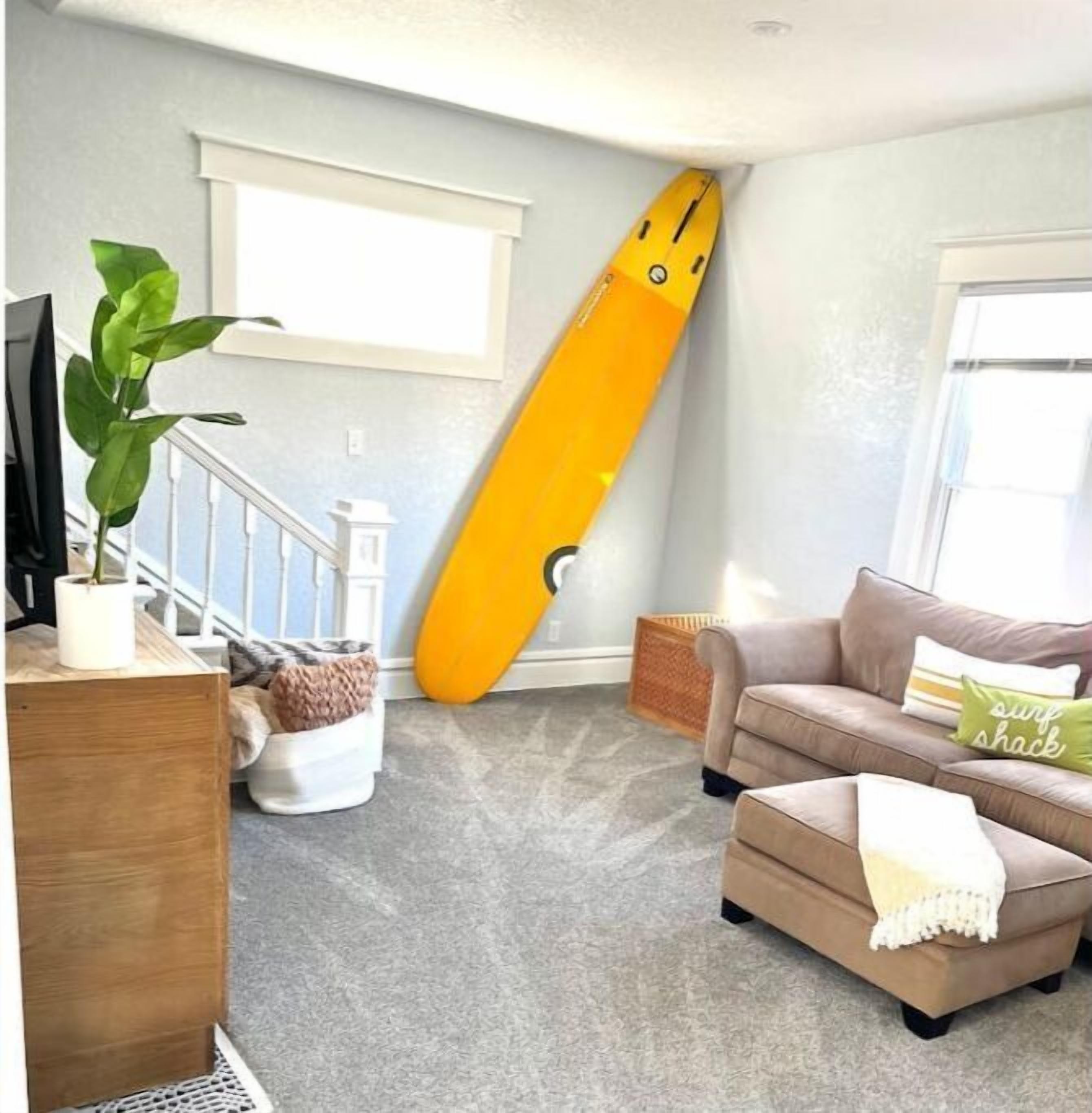 Ein Wohnzimmer mit großem Fenster, einer Treppe, einem gelben Surfbrett, das an die Wand gelehnt ist, einem beigen Sofa und einem Blumentopf.