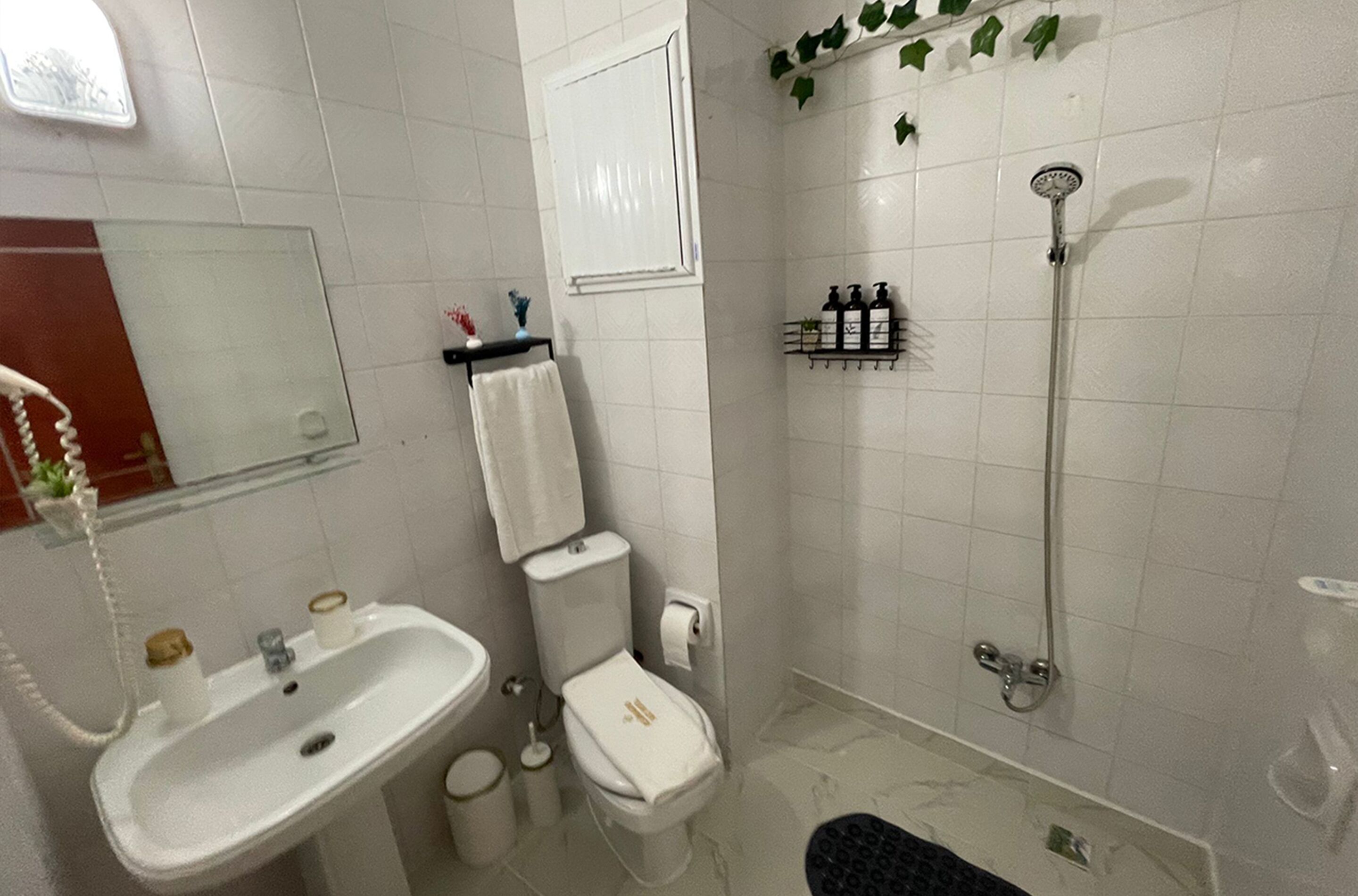 A bathroom with a sink, toilet, and shower. There is a mirror above the sink and a shelf with toiletries.