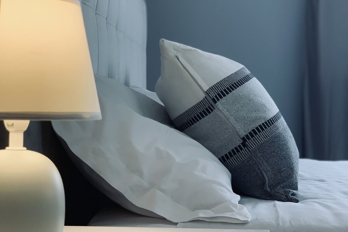 Ein ordentlich bezogenes Bett mit einem weißen und einem grauen Kissen mit Streifenmuster. Ein weißer Nachttisch mit einer Lampe ist zu sehen.
