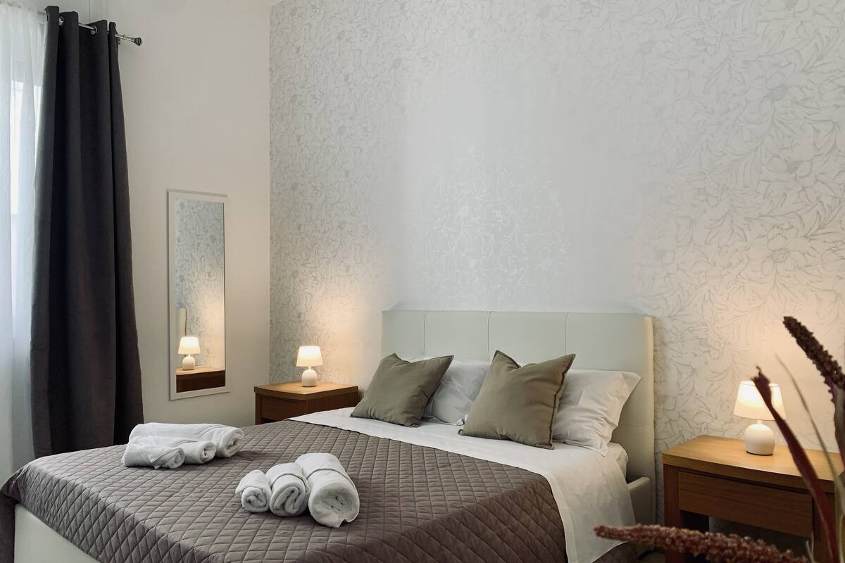 Ein ordentlich bezogenes Bett mit einer braunen Steude, zwei Nachttischlampen und einem floralen Wandpapier.