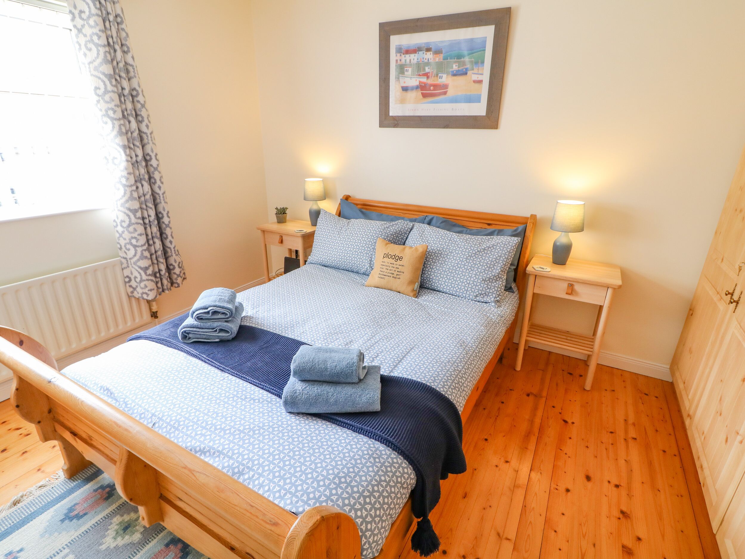A neatly made bed with a patterned duvet, two bedside lamps, a framed picture on the wall, and a window with curtains.