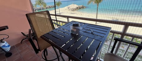 En terrasse med et bord og stole, udsigt over en strand med klart, blåt vand og palmer.