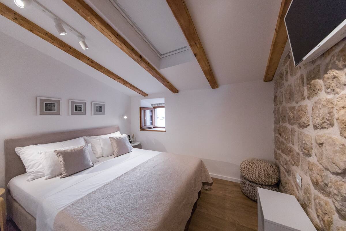 Une chambre avec un grand lit, des poutres en bois, un mur en pierre et une petite fenêtre.