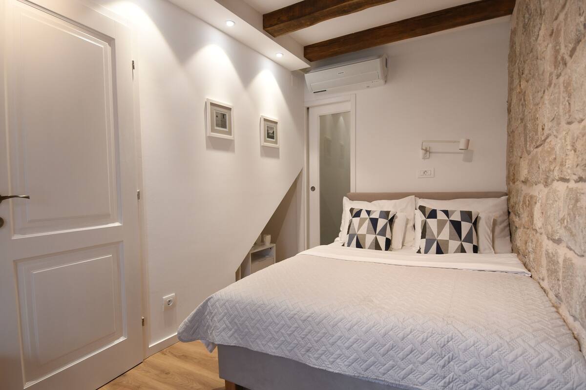 Une chambre à coucher avec un lit, des murs blancs, une poutre en bois au plafond, un mur en pierre et une porte.