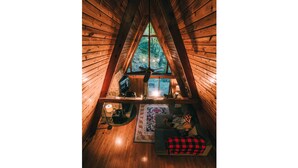 A cozy attic room with a wooden sloped ceiling, a ceiling fan, a television, and a view of the forest through a large window.
