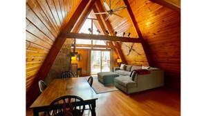 A cozy attic room with wooden walls and a sloped ceiling. There is a dining table with chairs, a sofa, and a small table with a lamp.