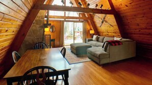 Un acogedor dormitorio en el ático con paredes de madera y techo inclinado. Hay una mesa de comedor con sillas, un sofá y una mesita de noche con una lámpara.